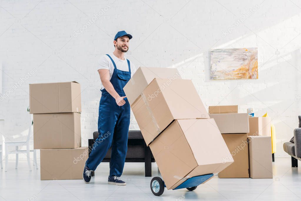 mover in uniform transporting  cardboard boxes on hand truck in apartment