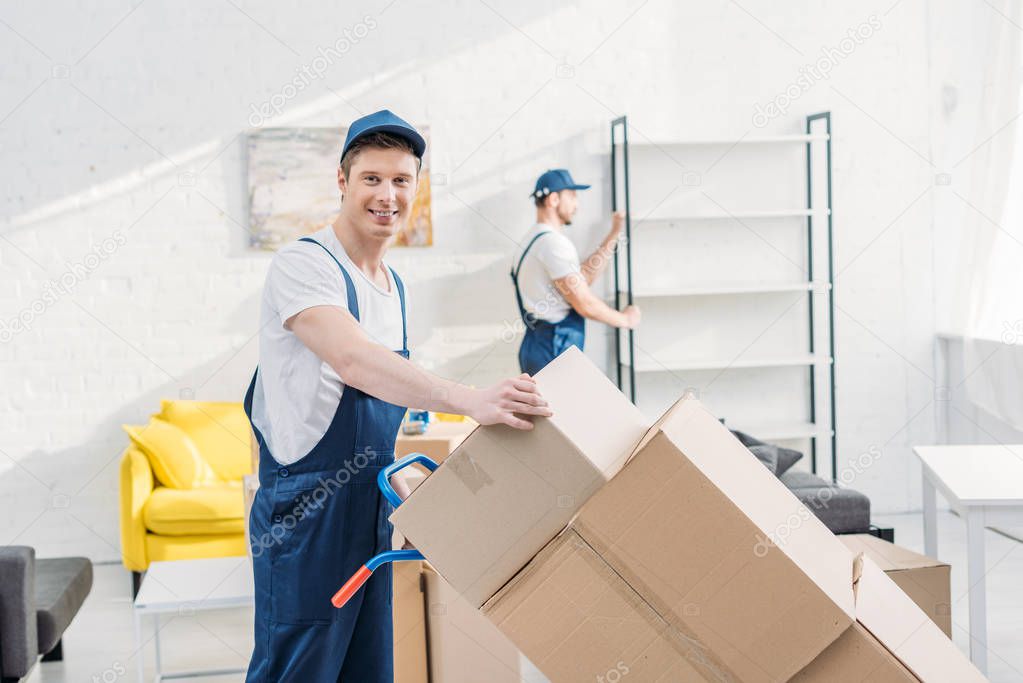 two movers in uniform transporting cardboard boxes and furniture in apartment