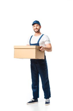 handsome mover in uniform carrying cardboard box, smiling and looking at camera isolated on white clipart