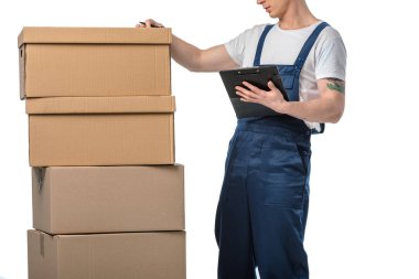 cropped view of mover in uniform holding clipboard near cardboard boxes isolated on white clipart