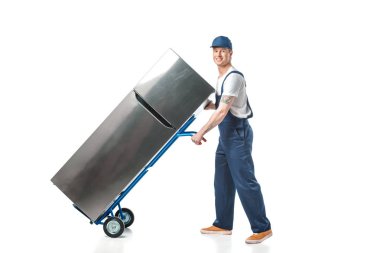 yakışıklı Mover üniformalı kamera bakıyor ve el kamyonunda buzdolabı taşıma beyaz izole