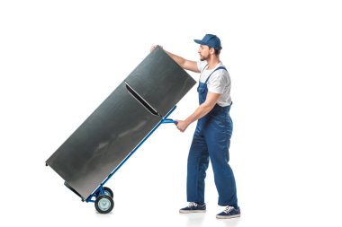 handsome mover in uniform transporting refrigerator on hand truck isolated on white clipart