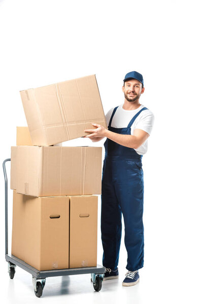 handsome mover in uniform transporting cardboard box near hand truck with packages isolated on white