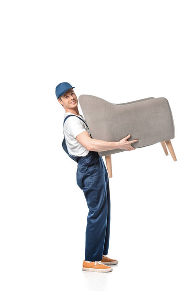 handsome mover in uniform carrying grey armchair, smiling and looking at camera isolated on white