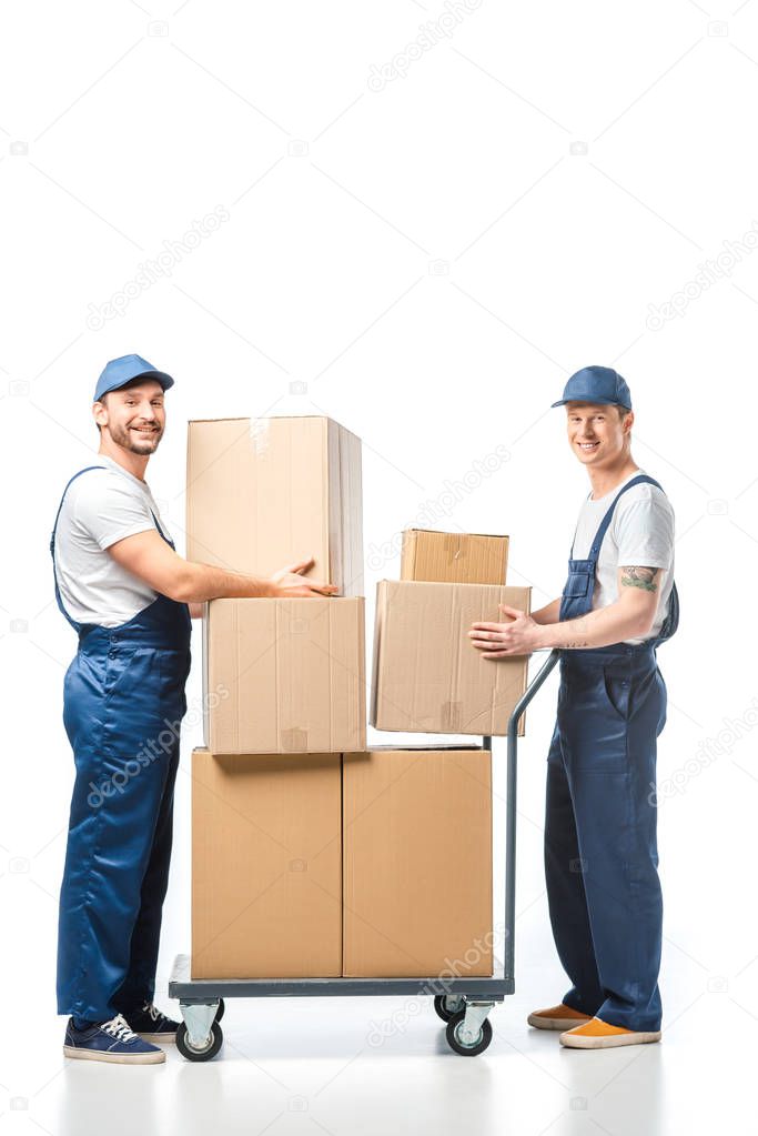 two handsome movers looking at camera while transporting cardboard boxes with hand truck on white