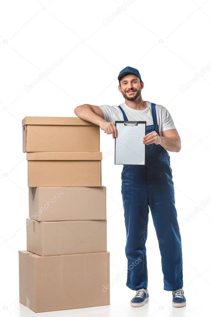 handsome mover in uniform looking at camera while holding blank clipboard near cardboard boxes isolated on white