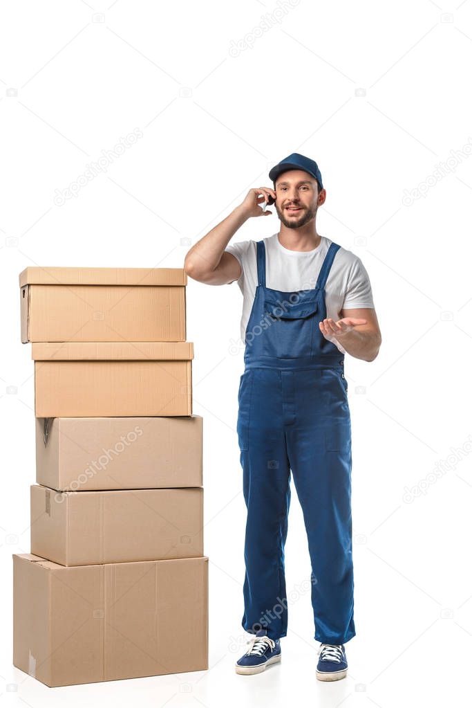 handsome smiling mover with cardboard boxes talking on smartphone and gesturing with hand isolated on white