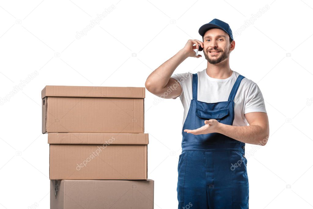 handsome smiling mover with cardboard boxes talking on smartphone and gesturing with hand isolated on white