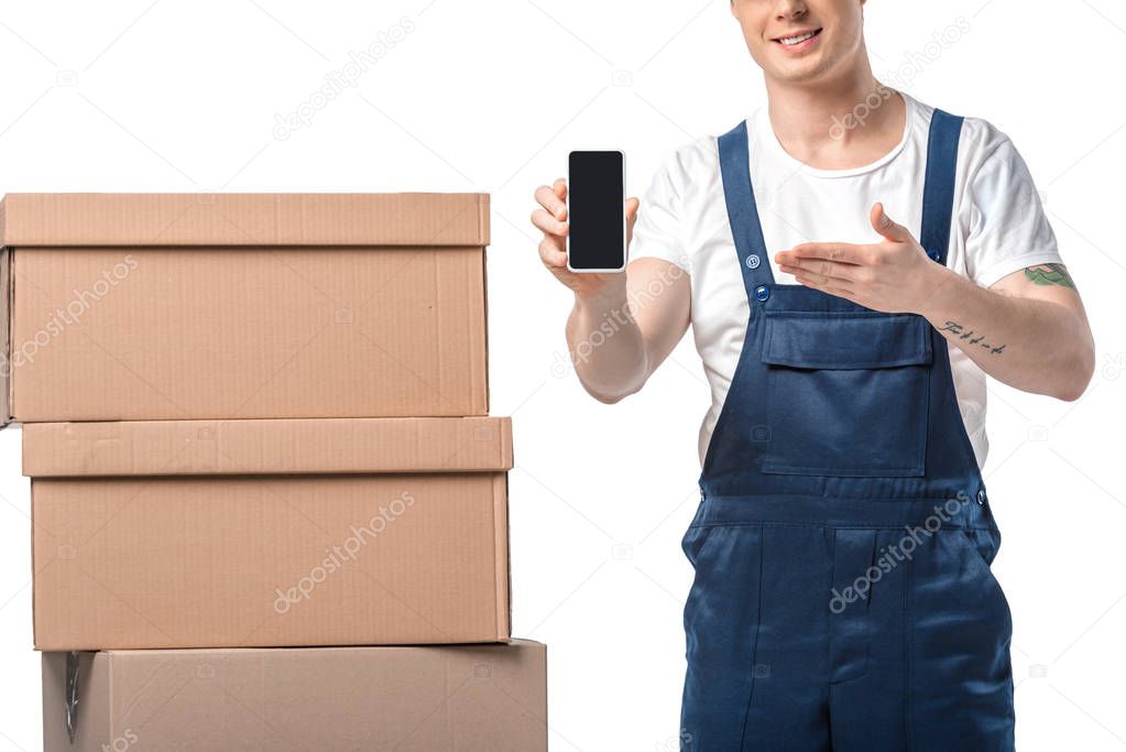 cropped view of smiling mover with cardboard boxes presenting smartphone with blank screen isolated on white