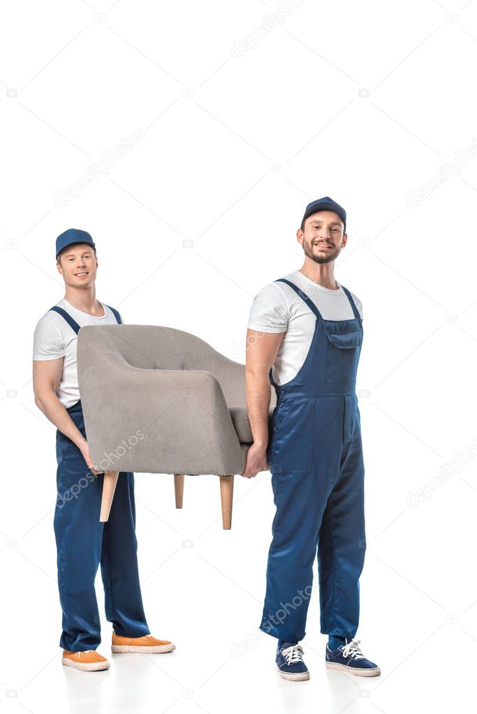 two smiling movers looking at camera and transporting grey armchair isolated on white