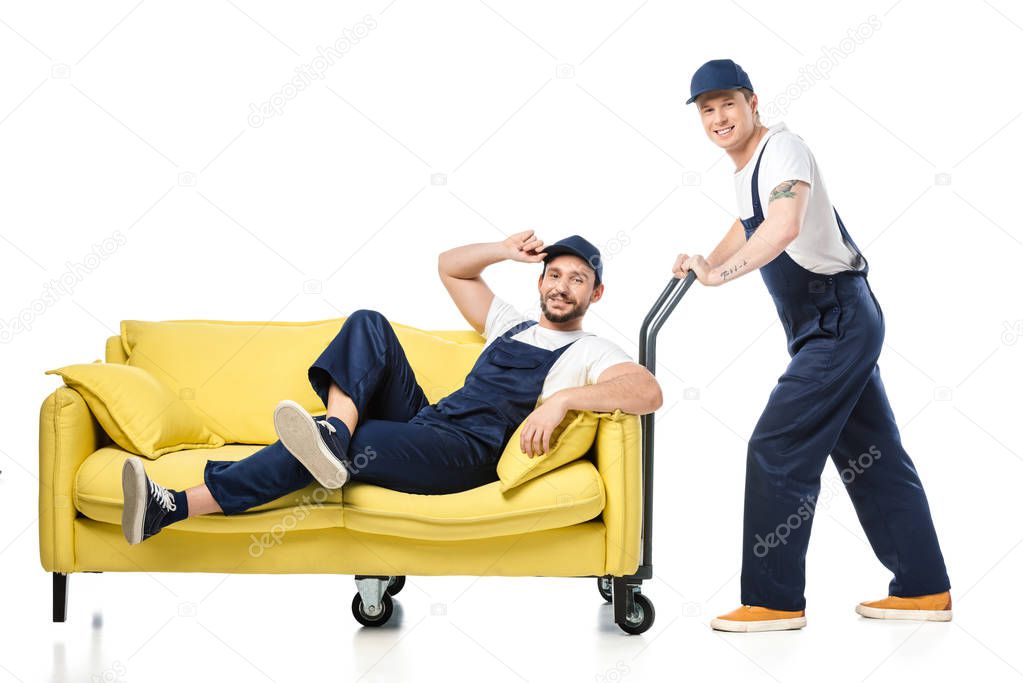 mover in uniform sitting on sofa and looking at camera while colleague pushing hand truck on white