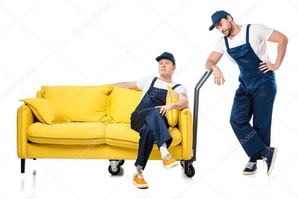 mover looking at colleague in uniform sitting on hand truck with sofa on white