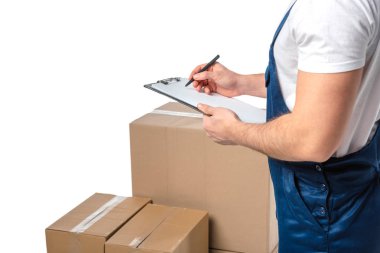 partial view of mover in uniform writing in clipboard near cardboard boxes isolated on white with copy space clipart