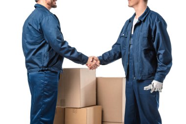 cropped view of two movers in uniform shaking hands near cardboard boxes isolated on white clipart