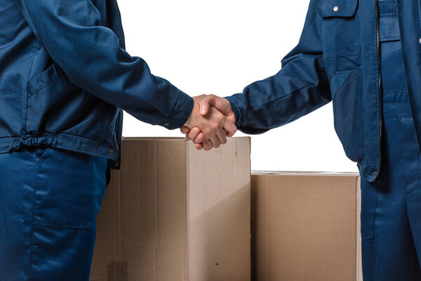 cropped view of two movers in uniform shaking hands near cardboard boxes isolated on white