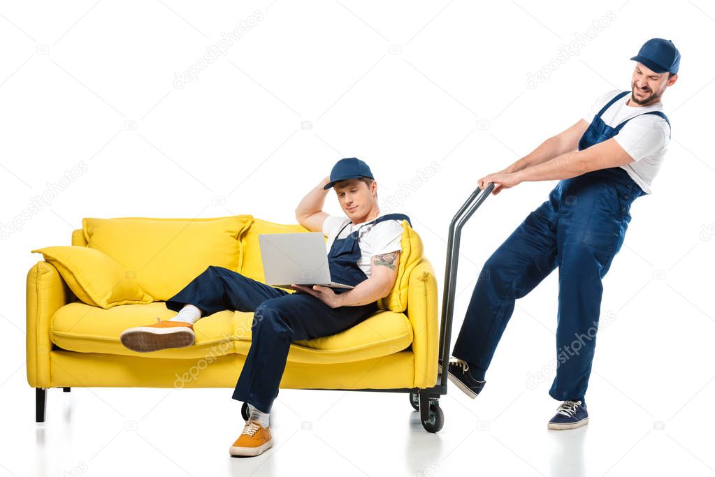 mover in uniform sitting on sofa and using laptop while colleague pushing hand truck on white