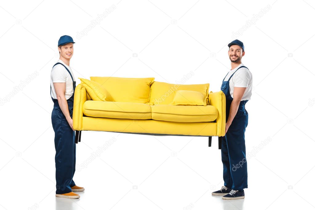 two smiling movers transporting yellow sofa and looking at camera on white