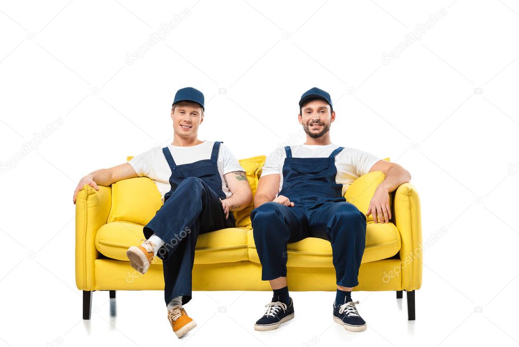 two smiling movers in uniform sitting on yellow sofa and looking at camera isolated on white