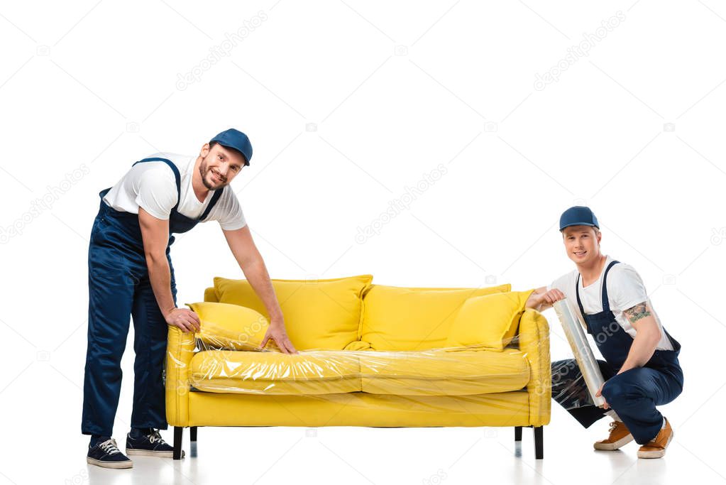 two movers looking at camera while wrapping yellow sofa with roll of stretch film on white