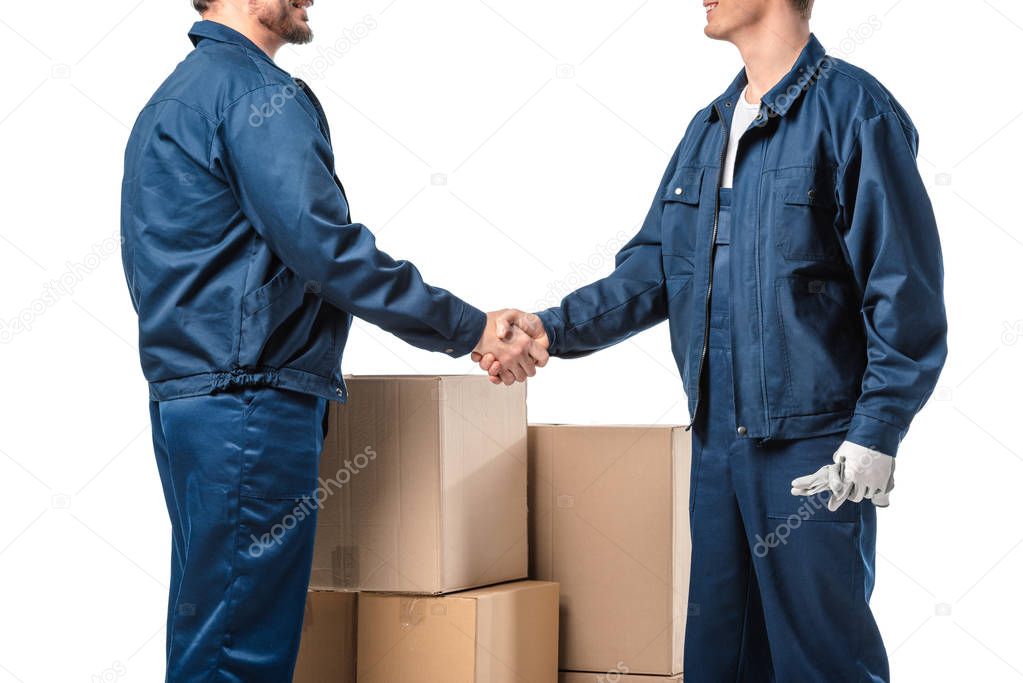 cropped view of two movers in uniform shaking hands near cardboard boxes isolated on white