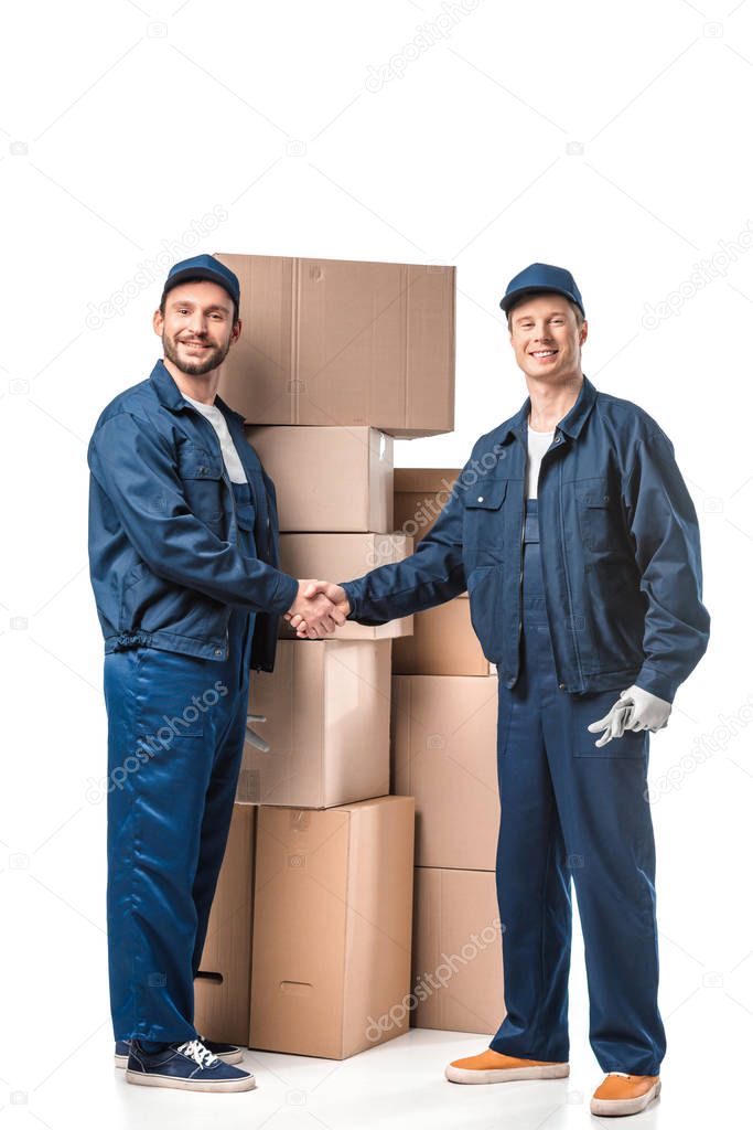two smiling movers in uniform looking at camera and shaking hands near cardboard boxes on white