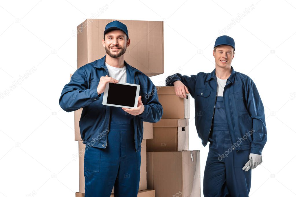 two movers with cardboard boxes looking at camera and presenting digital tablet with blank screen isolated on white