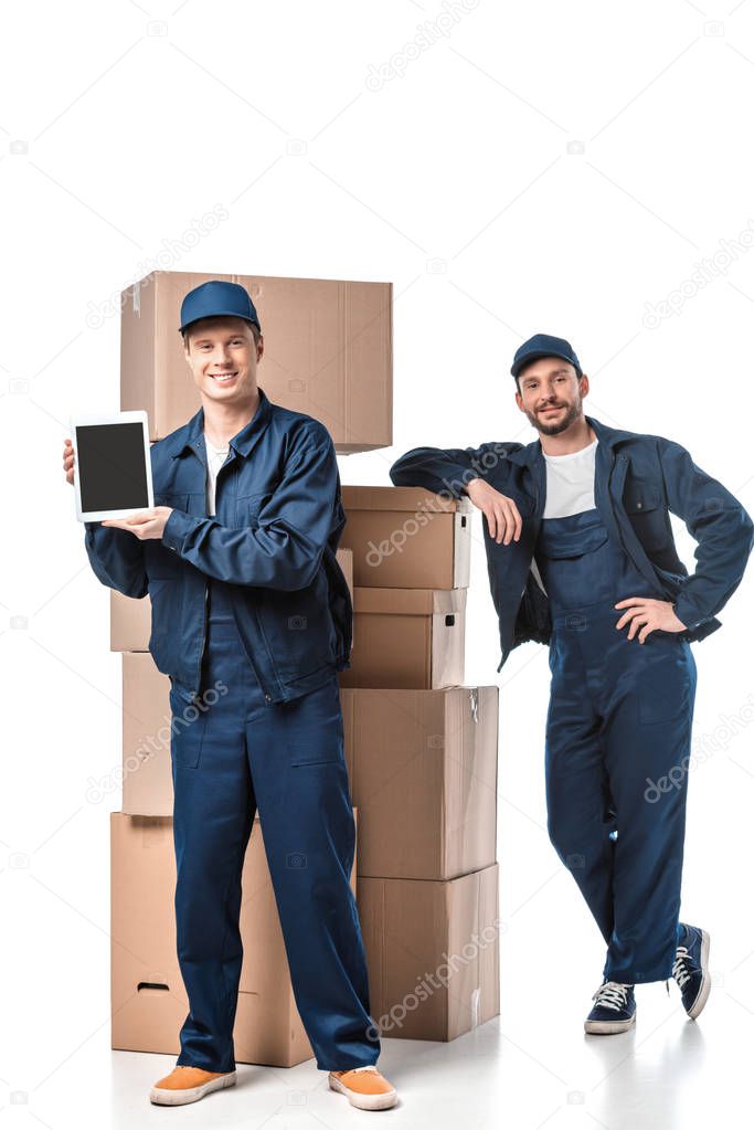 two movers with cardboard boxes looking at camera and presenting digital tablet with blank screen on white