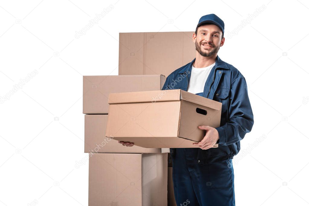 smiling handsome mover in uniform carrying cardboard box and looking at camera isolated on white with copy space