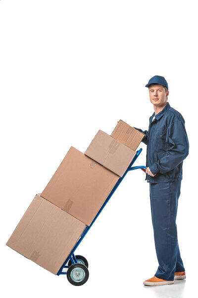 handsome mover in uniform looking at camera while transporting cardboard boxes on hand truck isolated on white