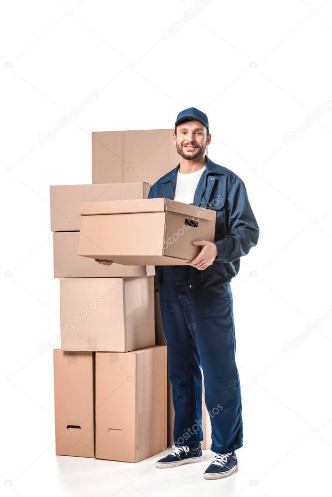 handsome mover in uniform carrying cardboard box, smiling and looking at camera isolated on white