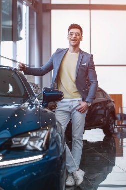 selective focus of cherful man in glasses standing near automobile with crossed legs in car showroom  clipart