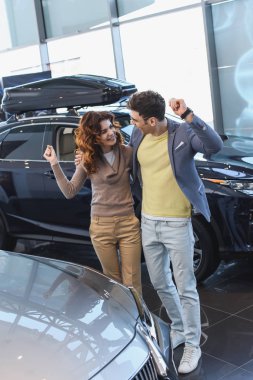 cheerful man in glasses hugging curly attractive woman while celebrating triumph in car showroom  clipart