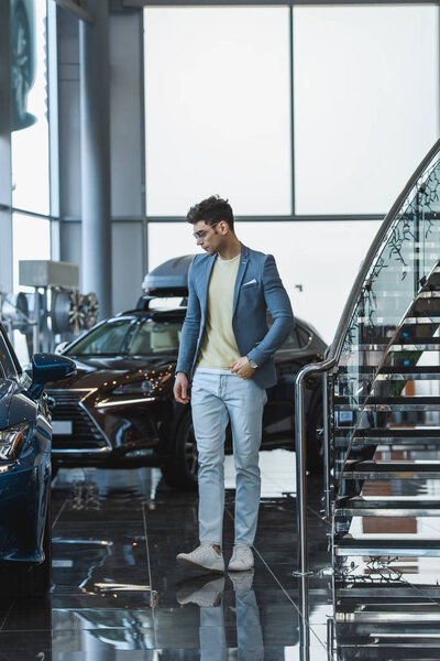 stylish man in glasses standing near automobiles in car showroom 