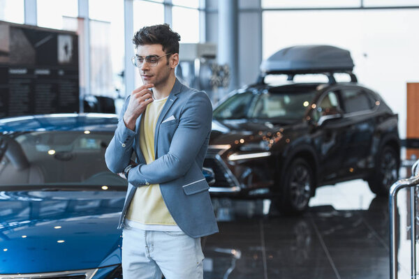 thoughtful man in glasses standing near automobiles in car showroom 
