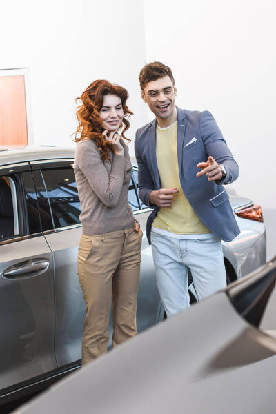 cheerful curly woman standing with happy man pointing with finger at car 