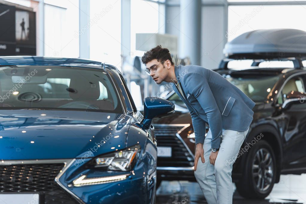 stylish man in glasses looking at automobile while standing in car showroom 
