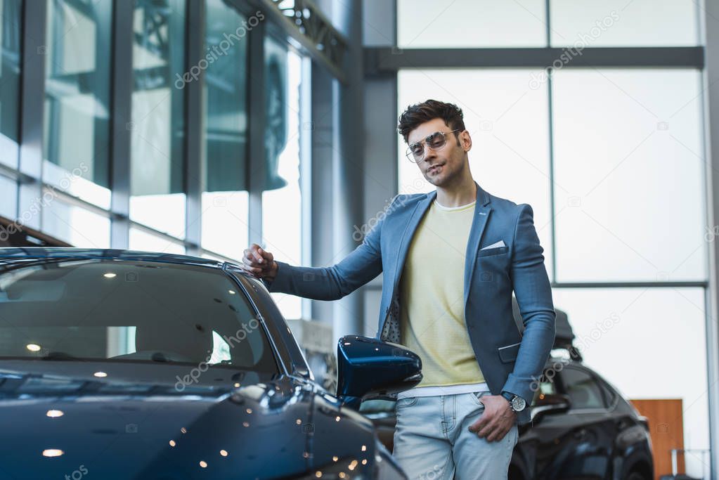 pensive man in glasses standing with hand in pocket near automobile in car showroom 