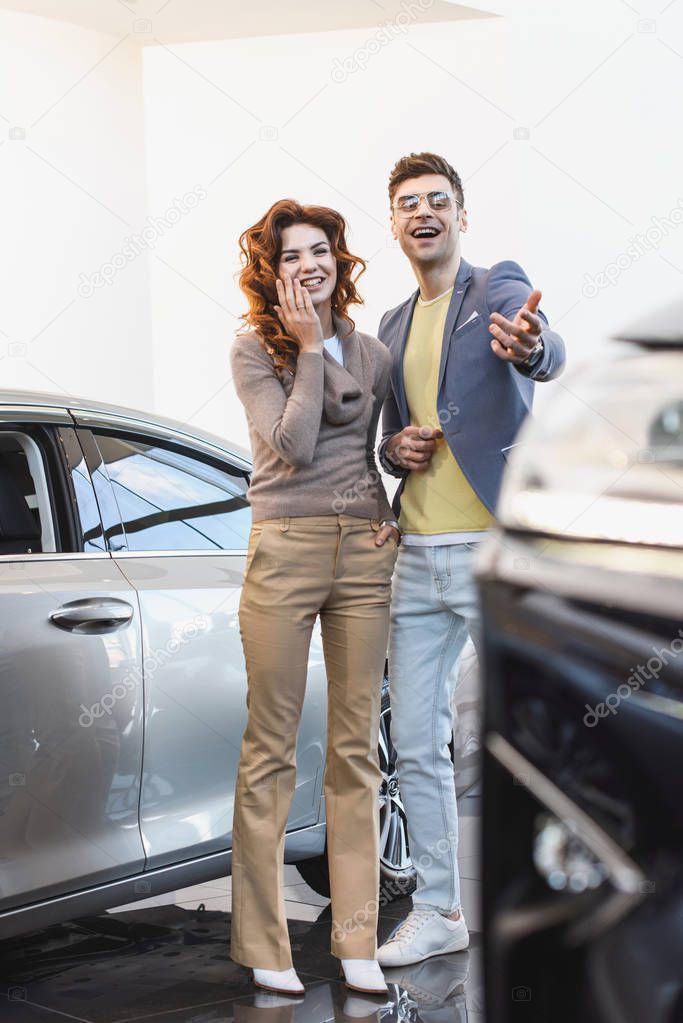 selective focus of cheerful curly woman standing with happy man pointing with finger at car 