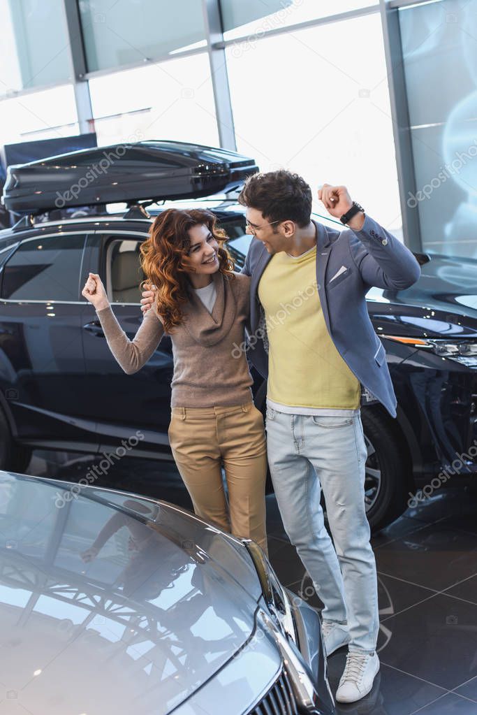 cheerful man in glasses hugging curly attractive woman while celebrating triumph in car showroom 