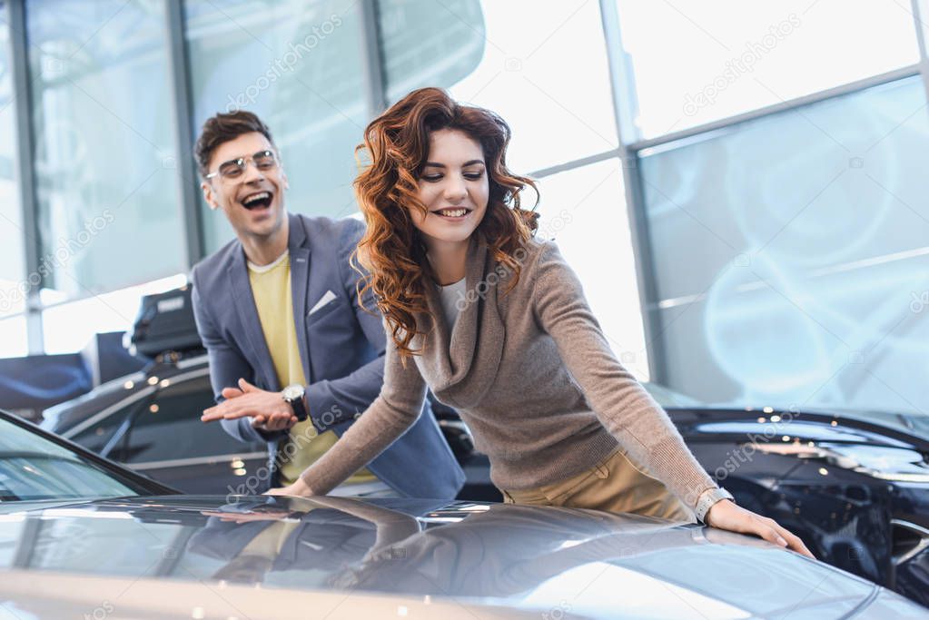 selective focus of happy curly woman smiling near excited man in glasses in car showroom