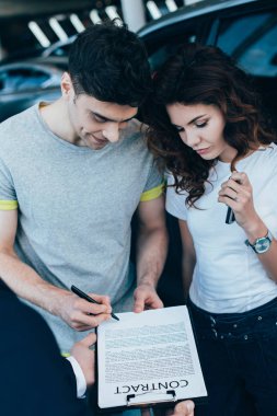 overhead view of car dealer holding clipboard while happy man signing contract near attractive curly woman   clipart