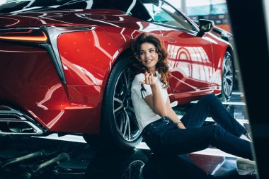 selective focus of happy woman sitting on floor near new luxurious car clipart