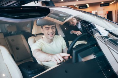 selective focus of handsome smiling man sitting in new car near car dealer clipart