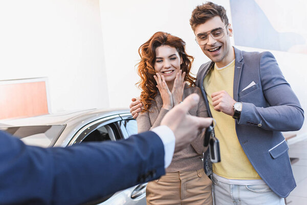 selective focus of cheerful man and woman looking at keys in hand of car dealer in car showroom 