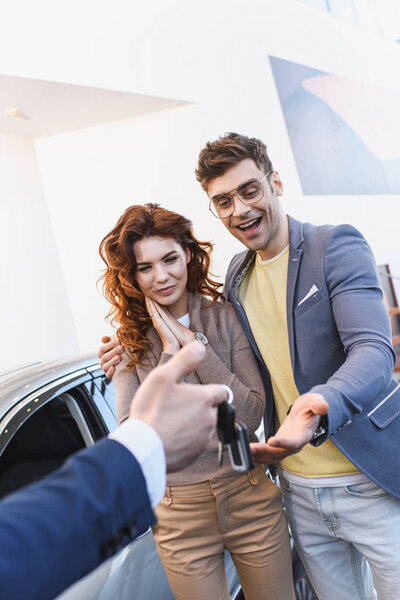 selective focus of happy man and woman looking at keys in hand of car dealer in car showroom 