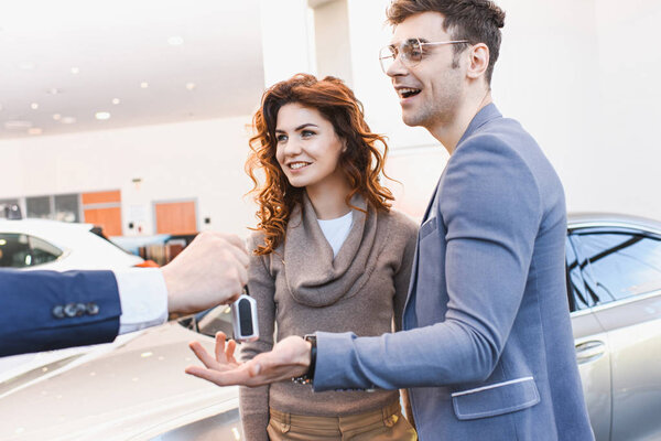 cropped view of car dealer holding keys near happy man in glasses and curly woman 