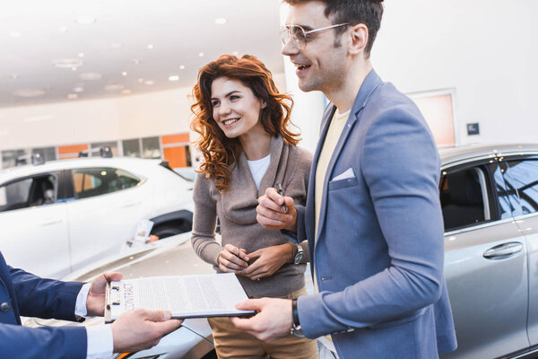 cropped view of car dealer holding clipboard with contract near happy man in glasses and curly woman 