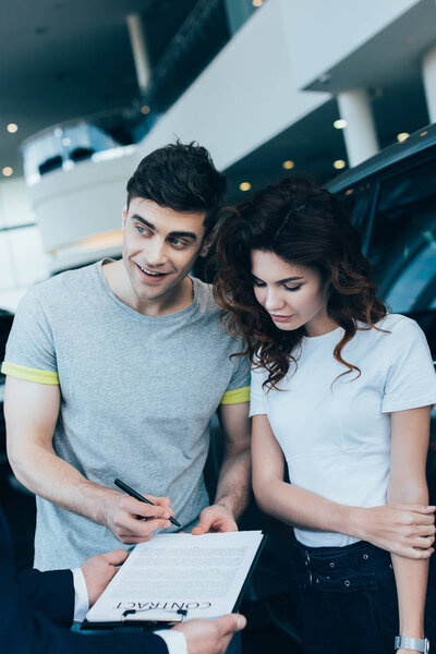 cropped view of car dealer holding clipboard with contract near happy man with pen and attractive woman  