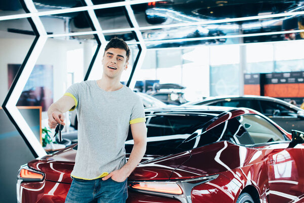 весёлый человек, стоящий с рукой в кармане и держащий ключи возле красной машины
 