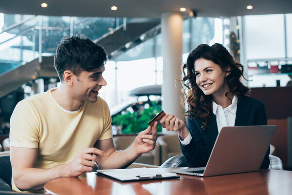 улыбающийся автомобильный дилер и клиент, сидящий за столом в автосалоне и держащий кредитную карту
